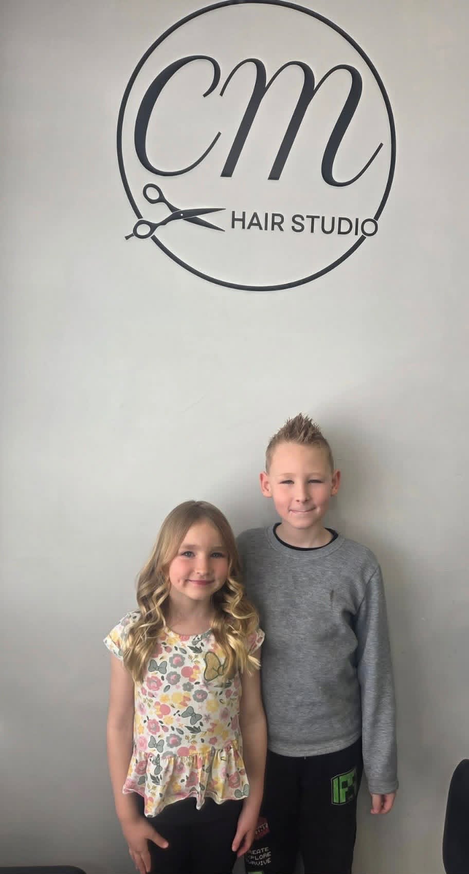 Two young clients with fresh cuts and styling in front of the CM Hair Studio logo wall